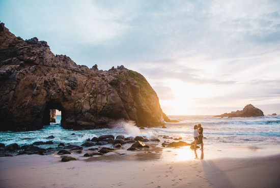 Engaged travel couple adventure to Pfeiffer Beach in Big Sur, California for photos at sunset next to the famous rock archway